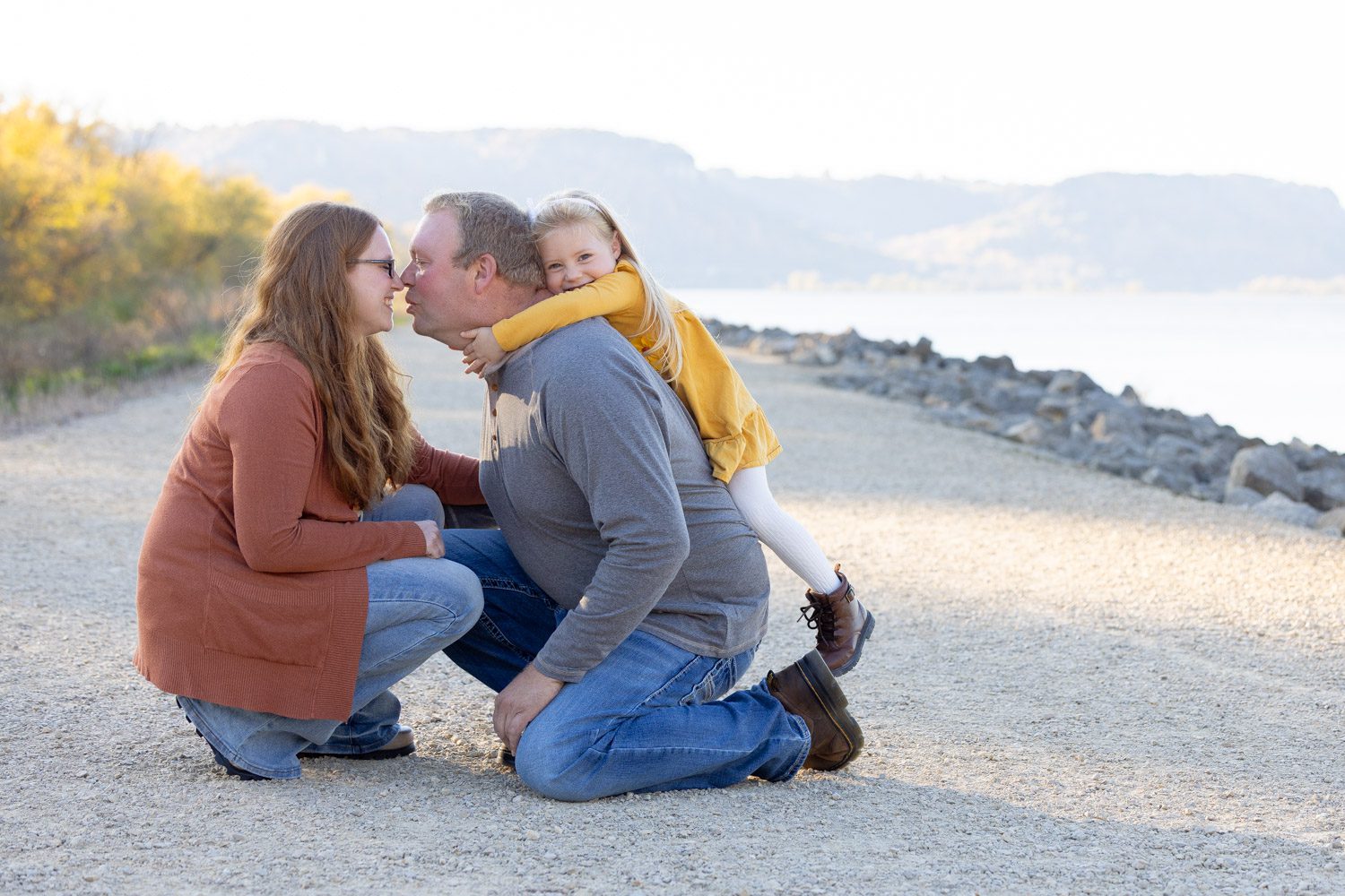 Mom and Dad are trying to sneak a kiss as their adorable little daughter climbs on Dad's back.